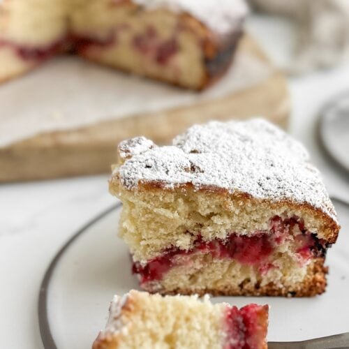 Close side view of a slice of cranberry vanilla coffee cake showing the cranberry ribbon layer, with a forkful of cake on the plate in front.