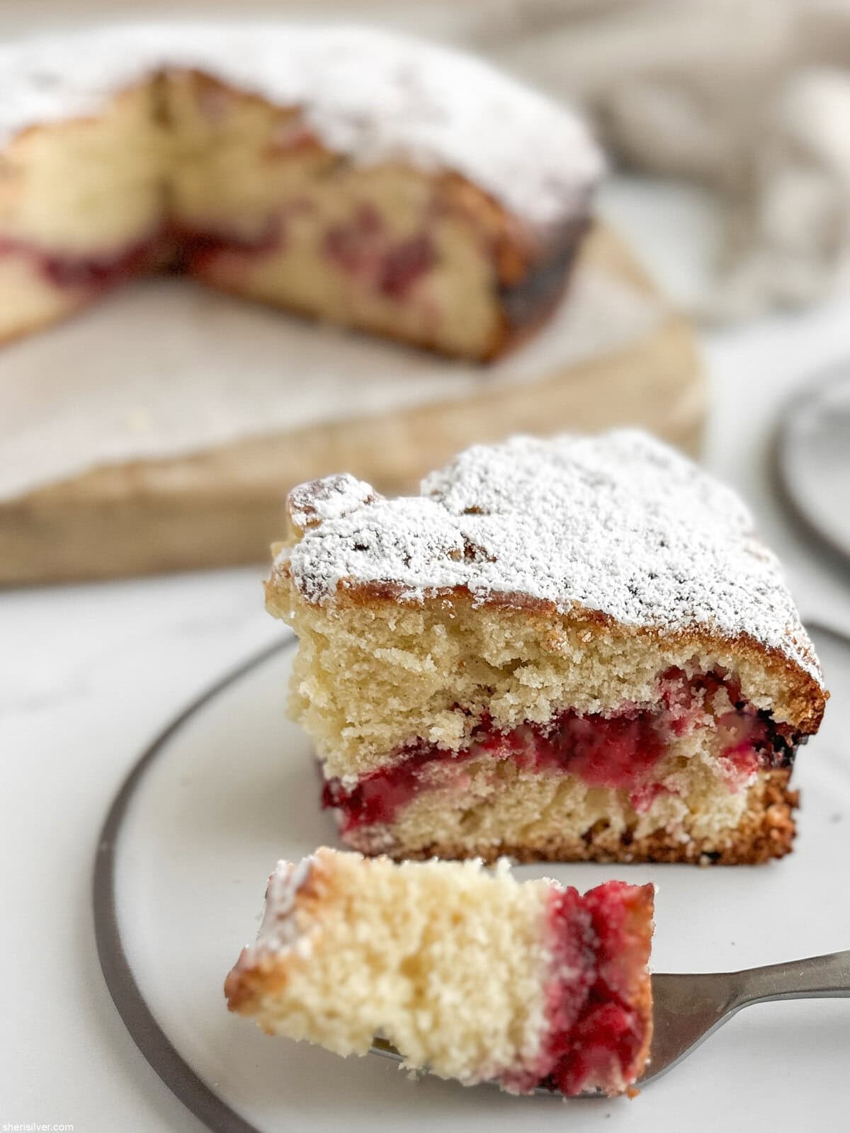 Close side view of a slice of cranberry vanilla coffee cake showing the cranberry ribbon layer, with a forkful of cake on the plate in front.