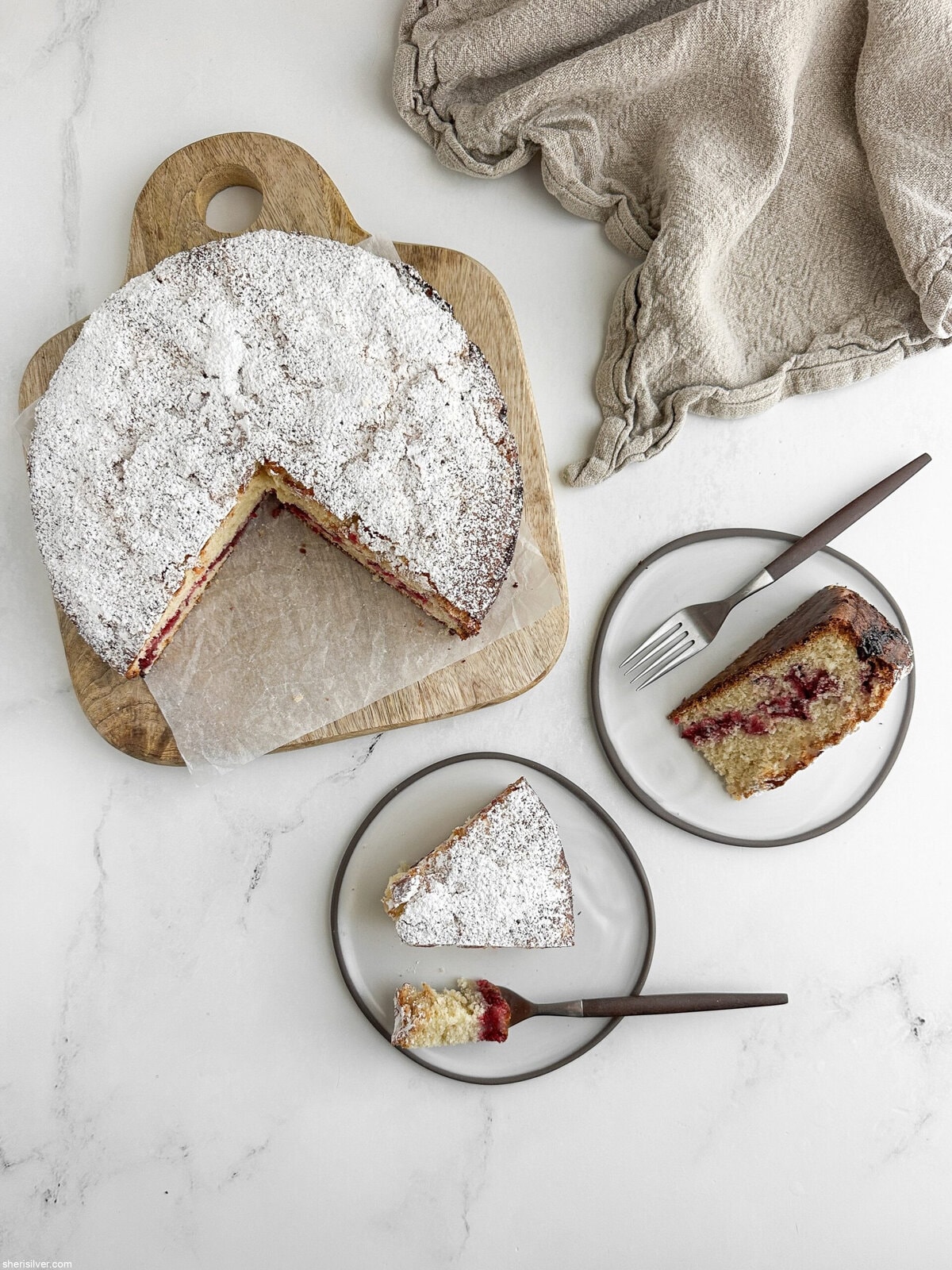 Overhead shot of a whole cranberry vanilla coffee cake on a wooden board with two plated slices nearby, ready for serving.
