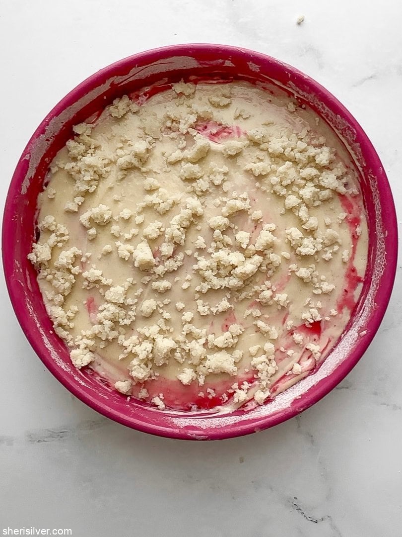 Round cake pan with layered batter, cranberries, and crumb topping ready for baking.