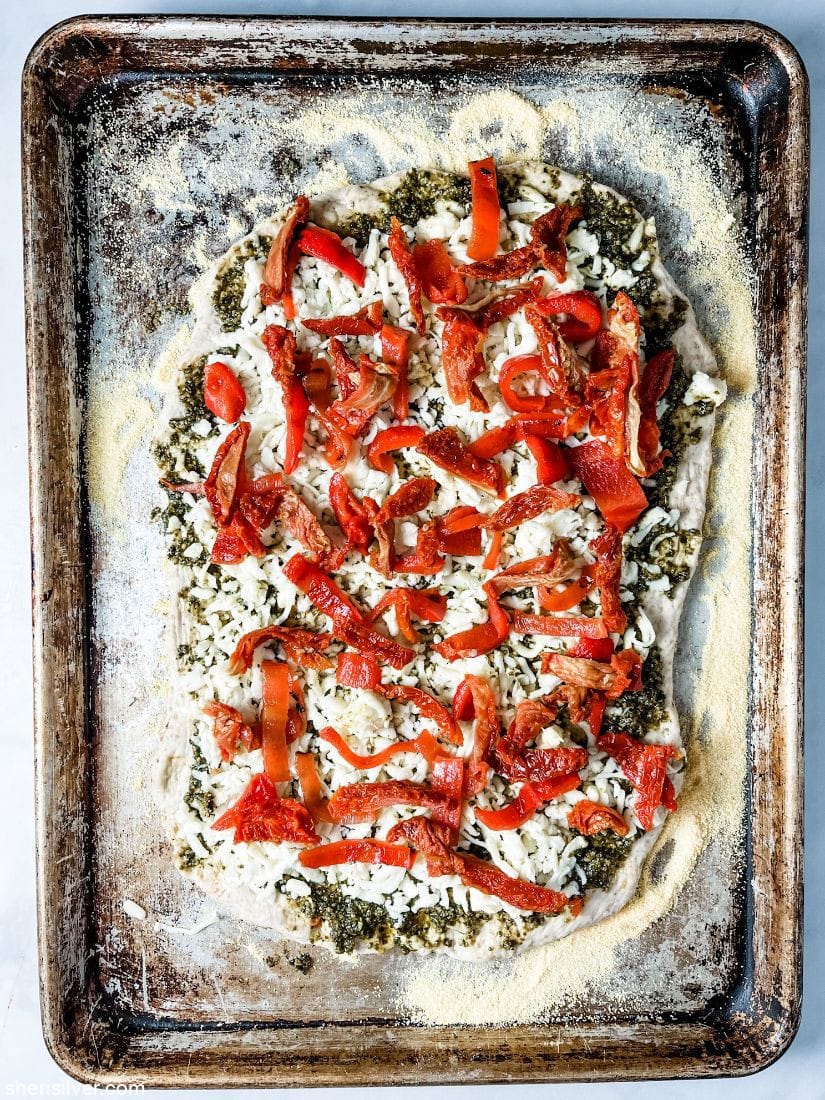 pizza dough on a sheet pan topped with pesto mozzarella and roasted peppers