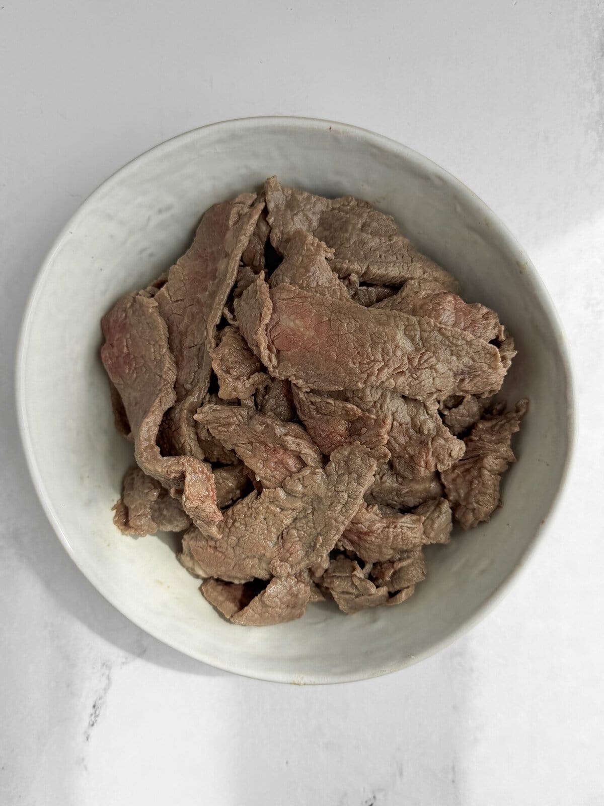 Blanched velveted flank steak strips in a white bowl — the beef will look slightly undercooked at this stage which is correct
