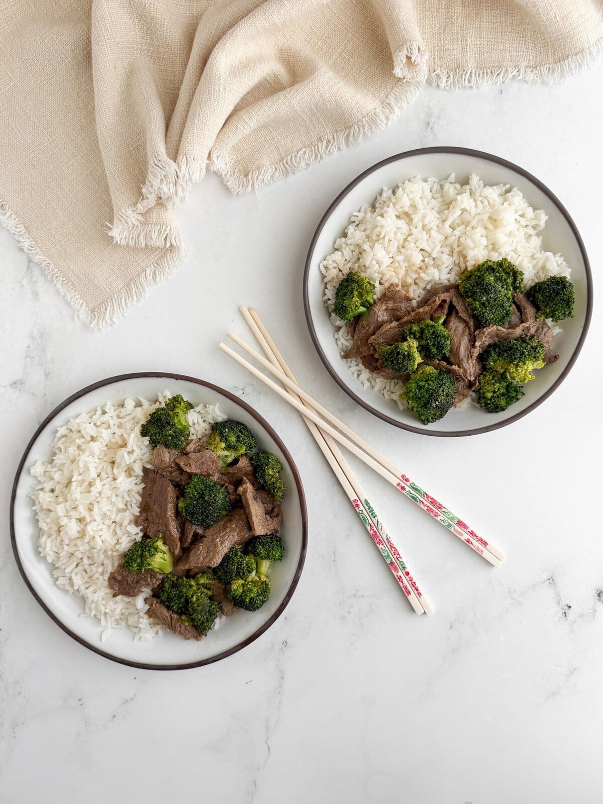 Overhead view of two bowls of velveted beef and broccoli with white rice, decorative chopsticks and a linen napkin on marble
