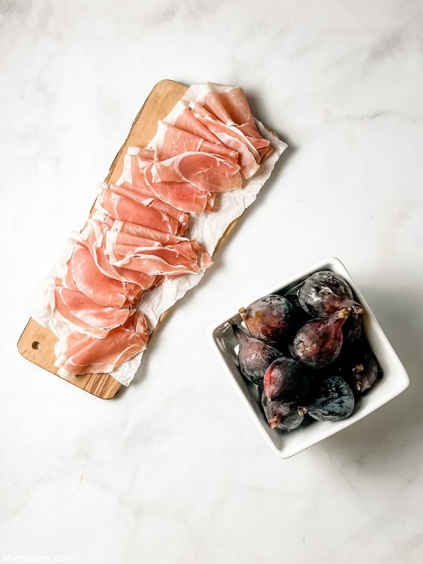 prosciutto on a wooden cutting board next to ceramic box of figs
