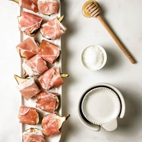 prosciutto wrapped figs on a white ceramic tray with honey stick and sea salt