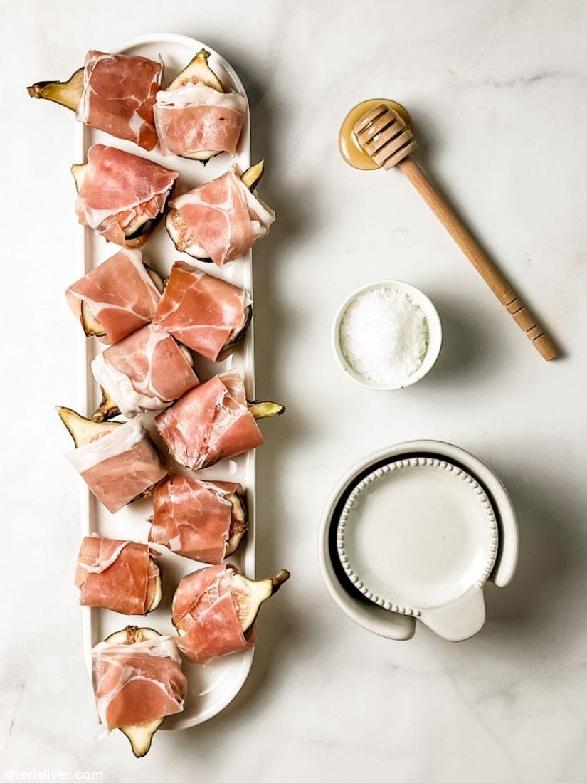 prosciutto wrapped figs on a white ceramic tray with honey stick and sea salt