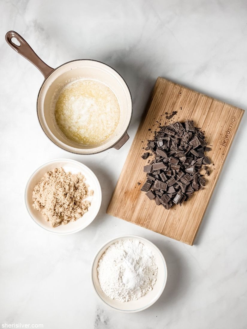 chopped chocolate on a cutting board next to bowls of brown sugar flour and melted butter