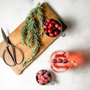 cocktail with sugared cranberry garnish next to tin cups filled with fresh and sugared cranberries and rosemary and scissors on a wooden board