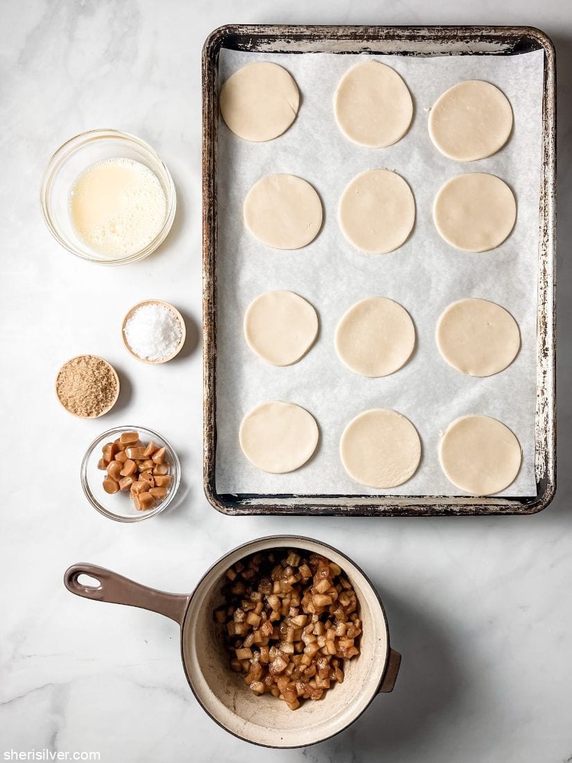 pie dough circles on a baking sheet next to bowls of caramels raw sugar flaky sea salt and a beaten egg