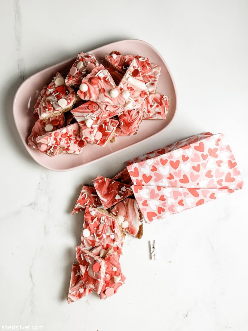 paper gift bag filled with valentine cookie bark next to a pink tray filled with valentine cookie bark