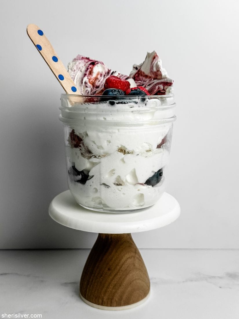 glass jar filled with berries whipped cream and meringue bark on a mini cake stand