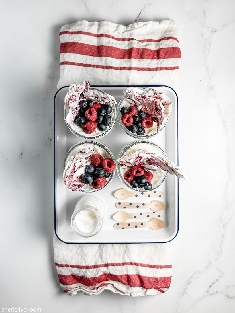 glass jars filled with berries whipped cream and meringue bark set in an enamel tray atop a red and white kitchen towel