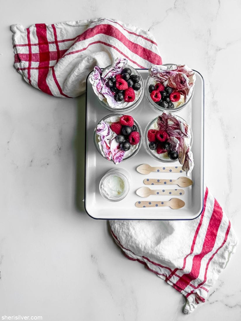 glass jars filled with berries whipped cream and meringue bark set in an enamel tray atop a red and white kitchen towel