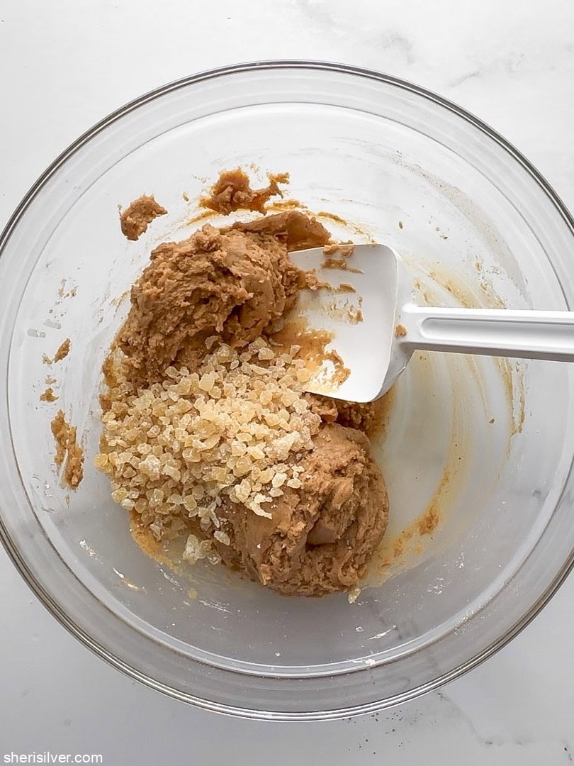 Pumpkin cookie dough in a glass bowl with chopped crystallized ginger being folded in with a spatula
