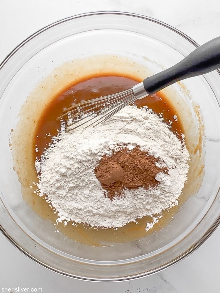 Dry ingredients like flour and pumpkin spice added to wet mixture in a glass bowl with a whisk