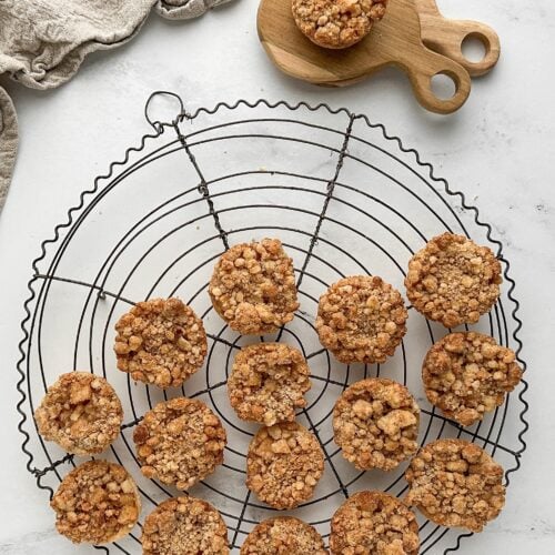 Apple streusel pie cookies cooling on a large round vintage wire rack, with one cookie styled on stacked mini wooden boards and a bit of natural linen.