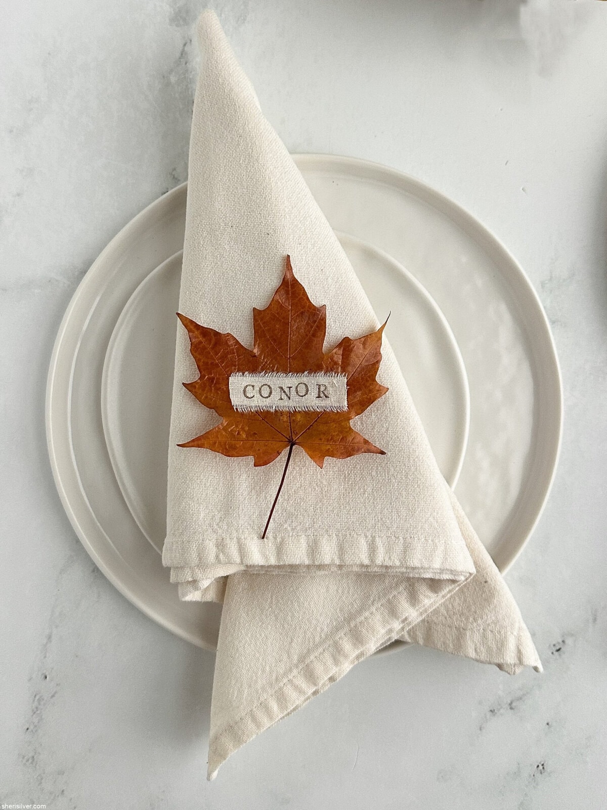 Thanksgiving table setting with DIY leaf place card on a napkin over white ceramic plates.