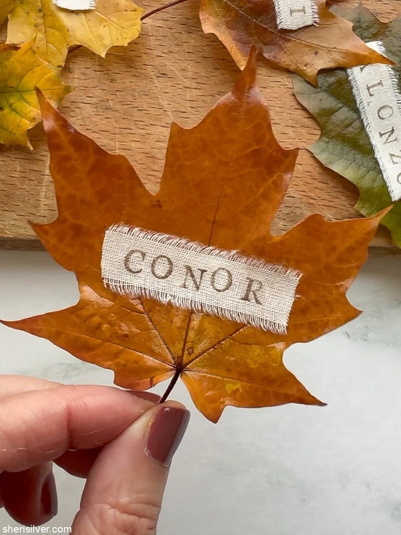 Close-up of a single DIY leaf place card with a stamped name ribbon attached to a preserved autumn leaf.