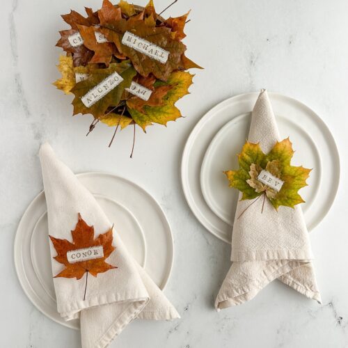 Thanksgiving table setting with DIY leaf place cards on napkins over white ceramic plates, next to a wooden bowl holding more place cards.