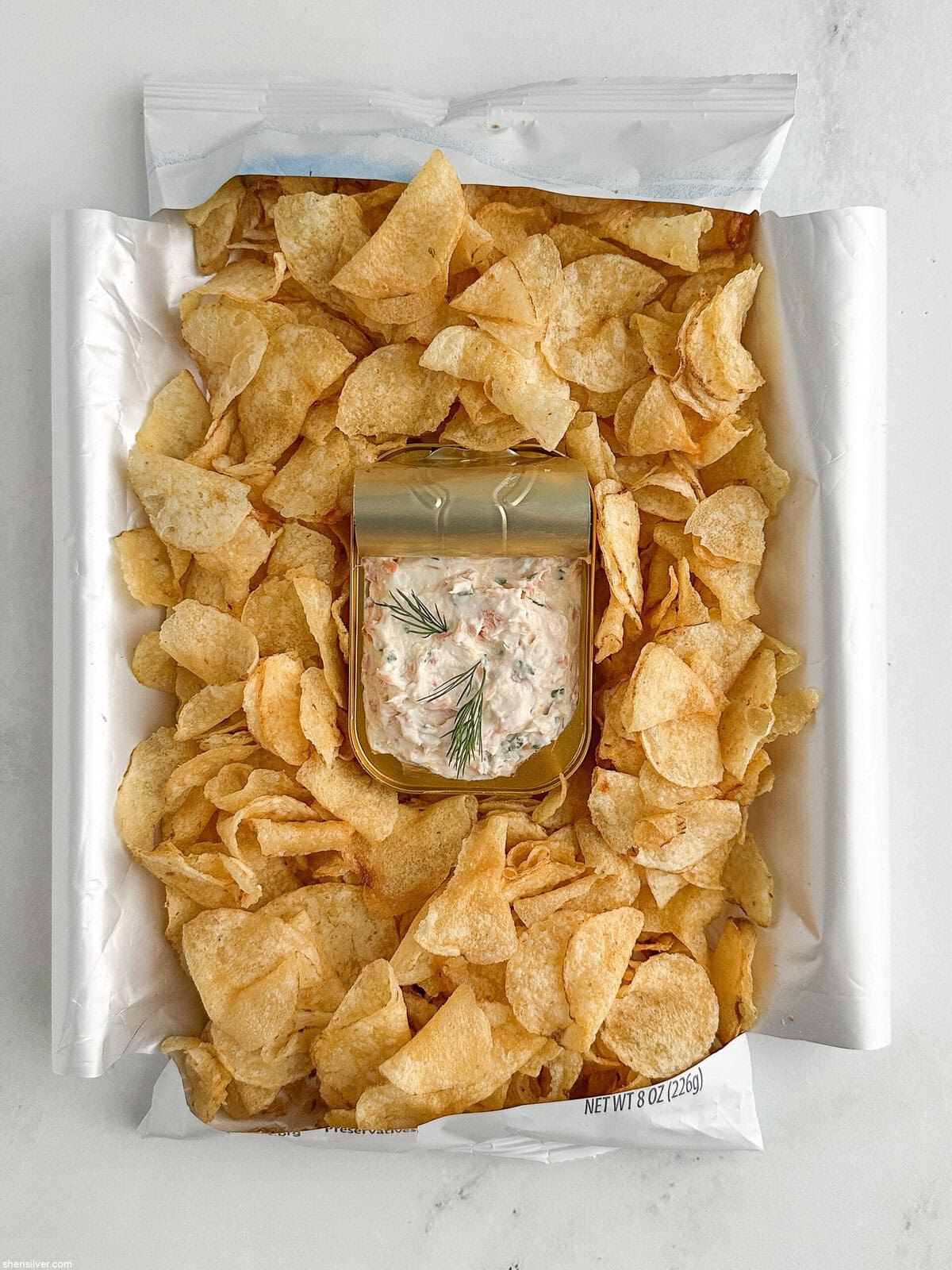 Overhead shot of a bag of potato chips laid on its back with sides opened like a tray, tin of dip nestled in the center for serving.