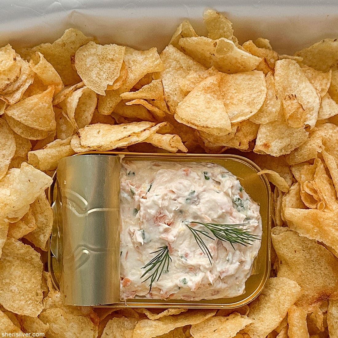 Overhead shot of a bag of potato chips laid on its back with sides opened like a tray, tin of dip nestled in the center for serving.