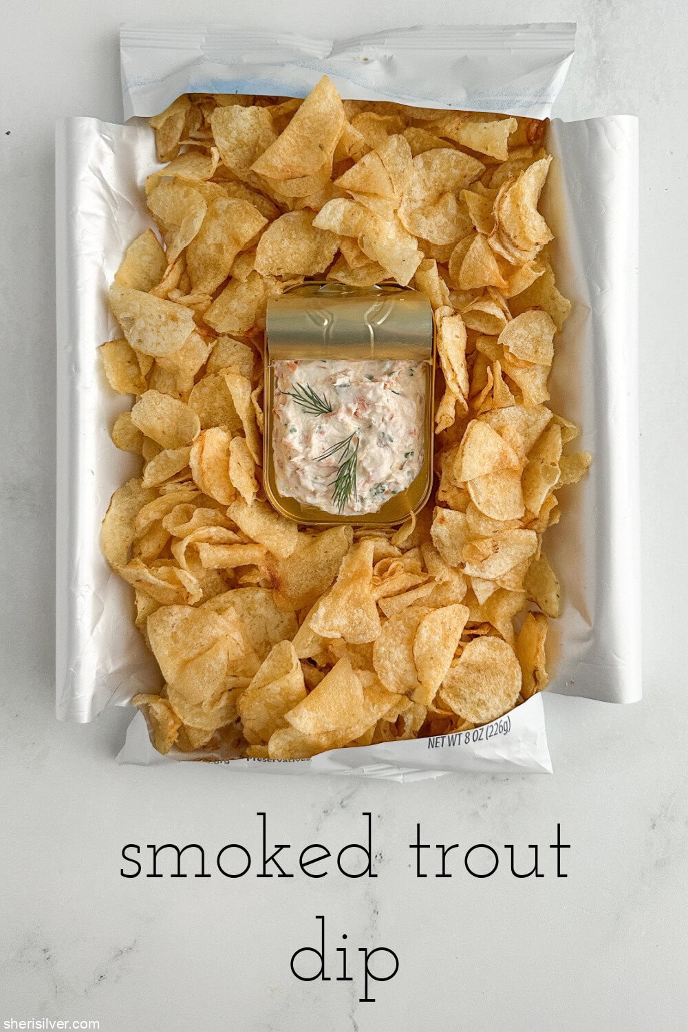 Overhead shot of a bag of potato chips laid on its back with sides opened like a tray, tin of dip nestled in the center for serving, with text overlay.