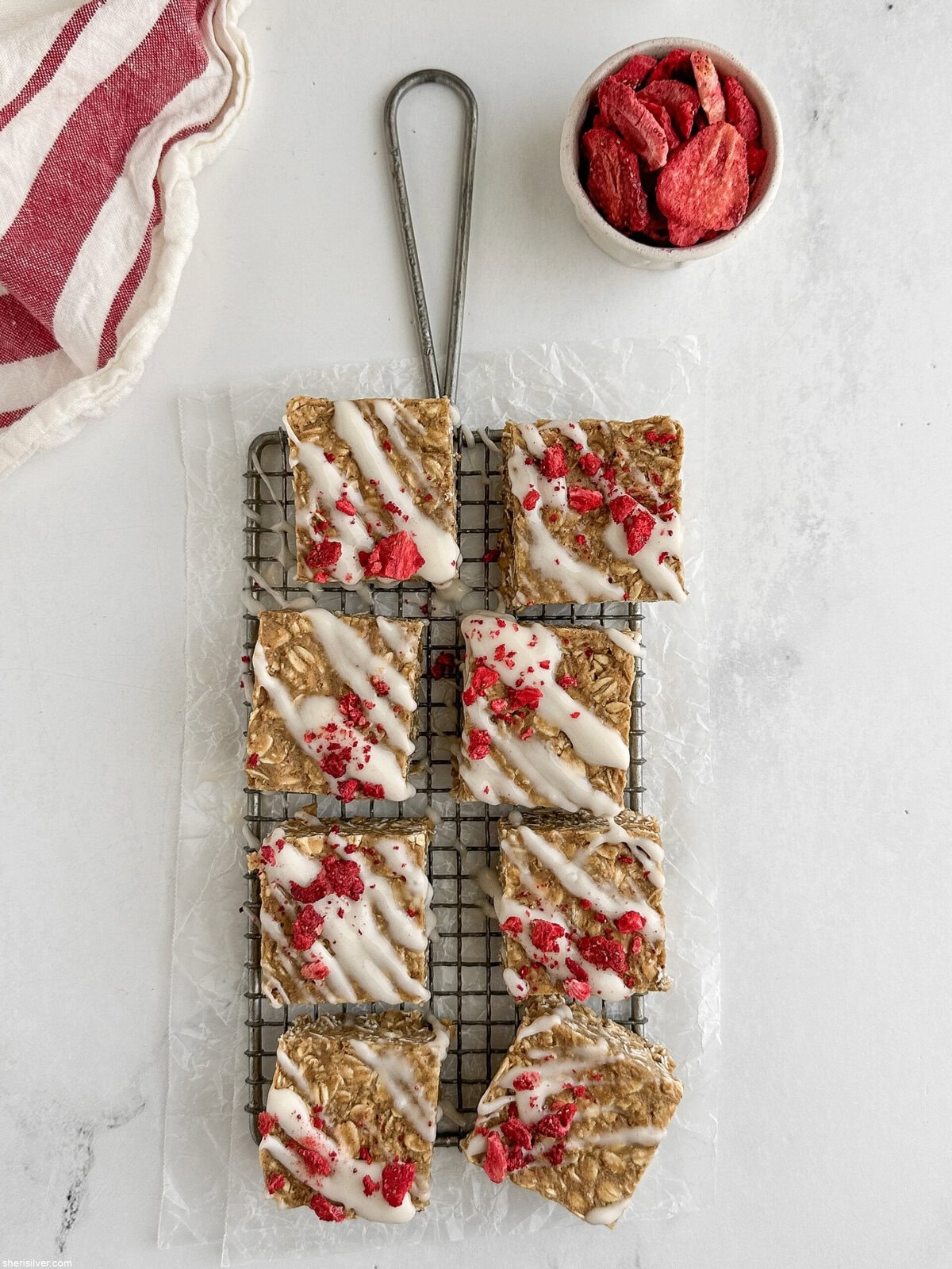 Overhead shot of finished vanilla tahini protein oat bars on a wire rack with a small ceramic dish of freeze-dried strawberries and a red-and-white linen towel.