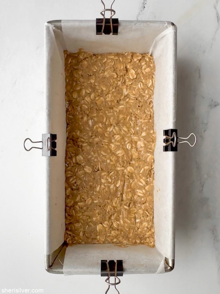 Vanilla tahini protein oat bar mixture pressed evenly into a parchment-lined 8×4 loaf pan, ready to chill — no-bake protein bars in progress.