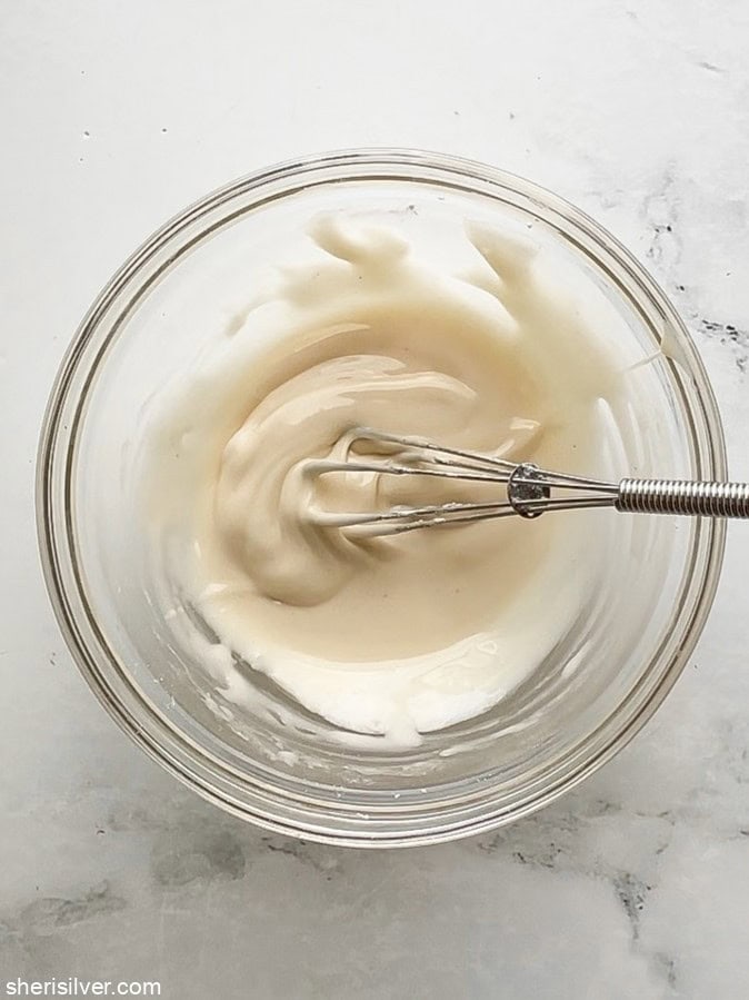 Overhead shot of a small glass bowl with vanilla glaze and a small metal whisk, ready for drizzling on oat bars.