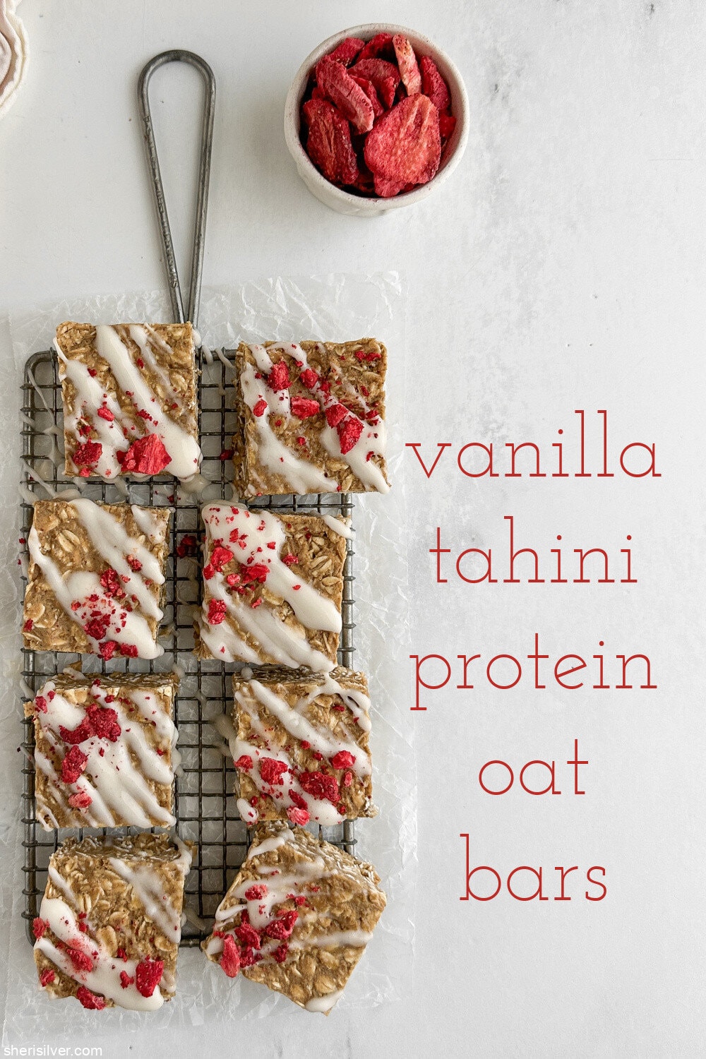 Overhead shot of finished vanilla tahini protein oat bars on a wire rack with a small ceramic dish of freeze-dried strawberries and a red-and-white linen towel.