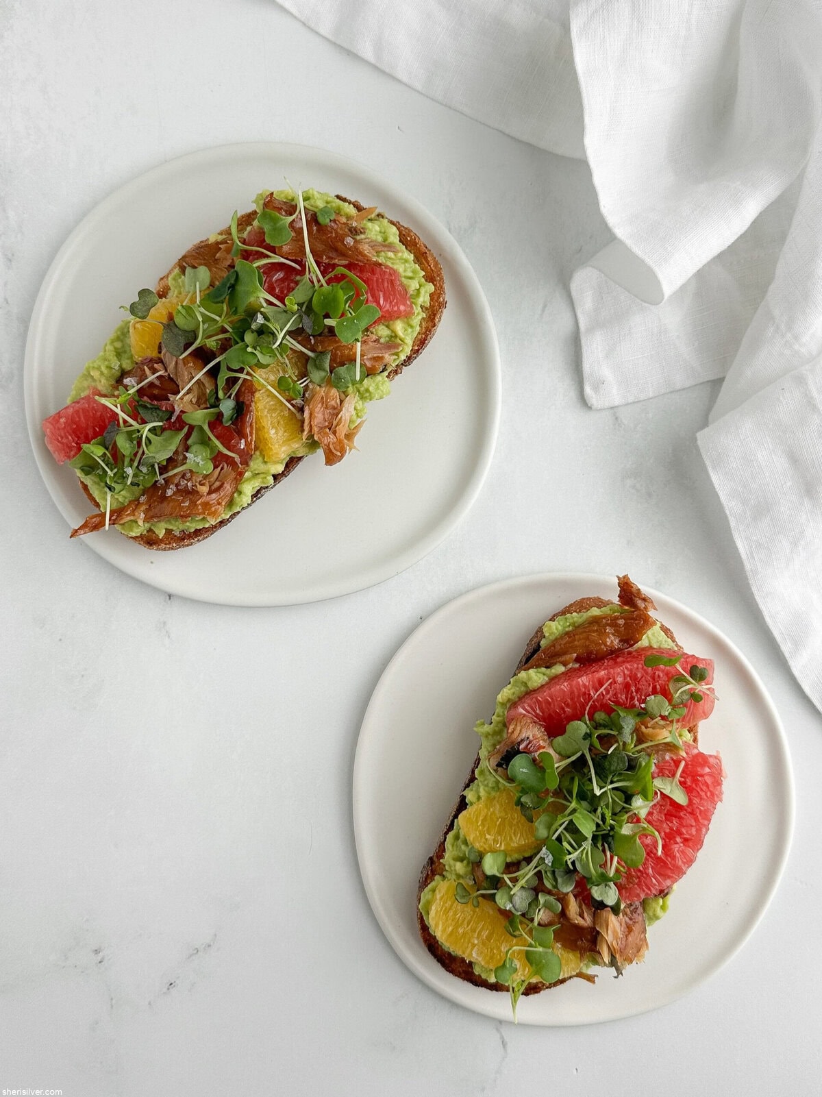Overhead view of smoked mackerel avocado toast on white plates with a white linen napkin.