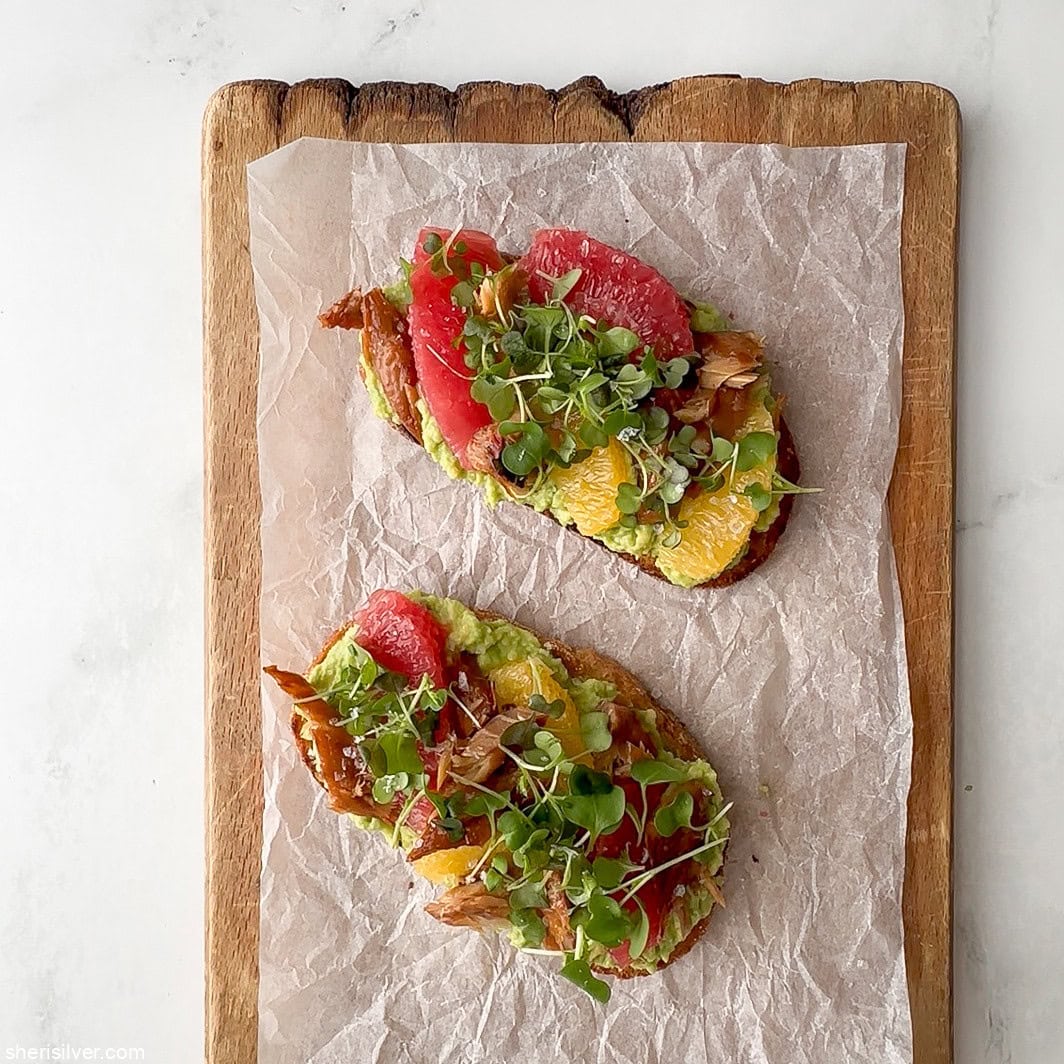 Smoked mackerel avocado toast topped with citrus and microgreens on a parchment-lined wooden board.