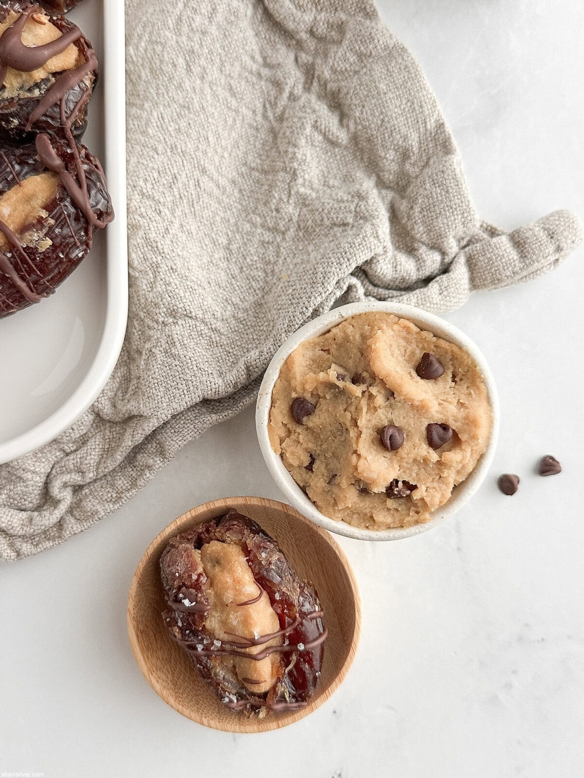 Overhead close-up of one cookie dough–stuffed date next to a small dish of cookie dough on a linen napkin.