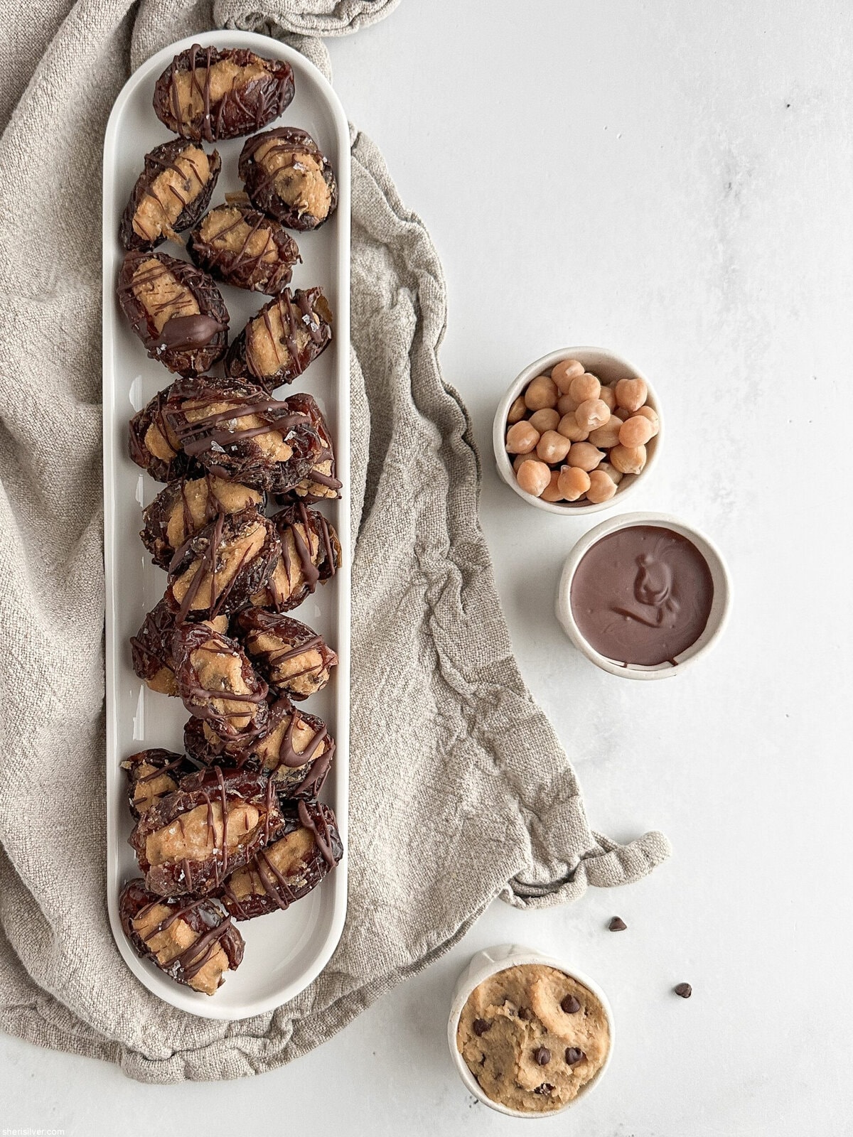 Overhead shot of finished stuffed dates on a white tray with small dishes of cookie dough, melted chocolate, and chickpeas on a linen napkin.