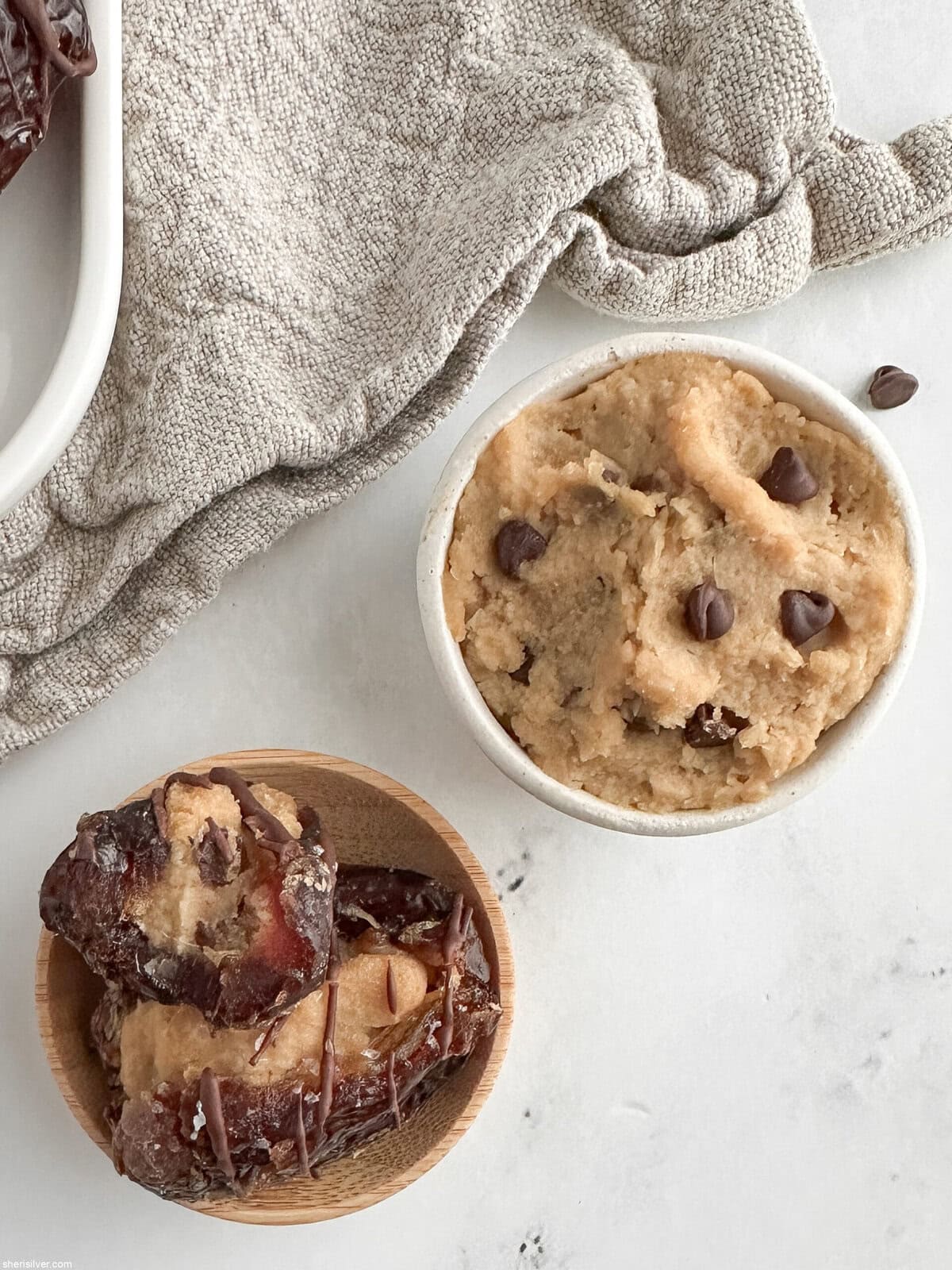 Close-up of a stuffed date with a bite taken out, next to a small dish of cookie dough on a linen napkin.
