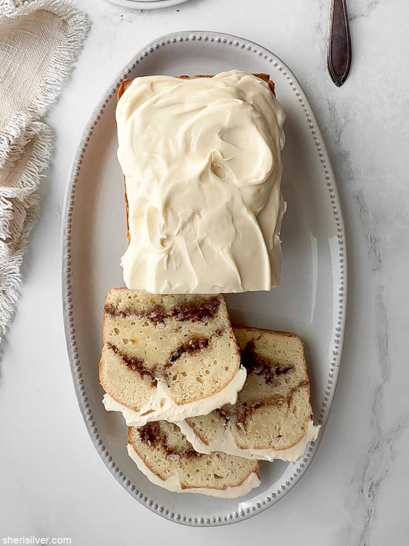 Finished cinnamon roll loaf cake, partially sliced on a white oval platter with a linen napkin.