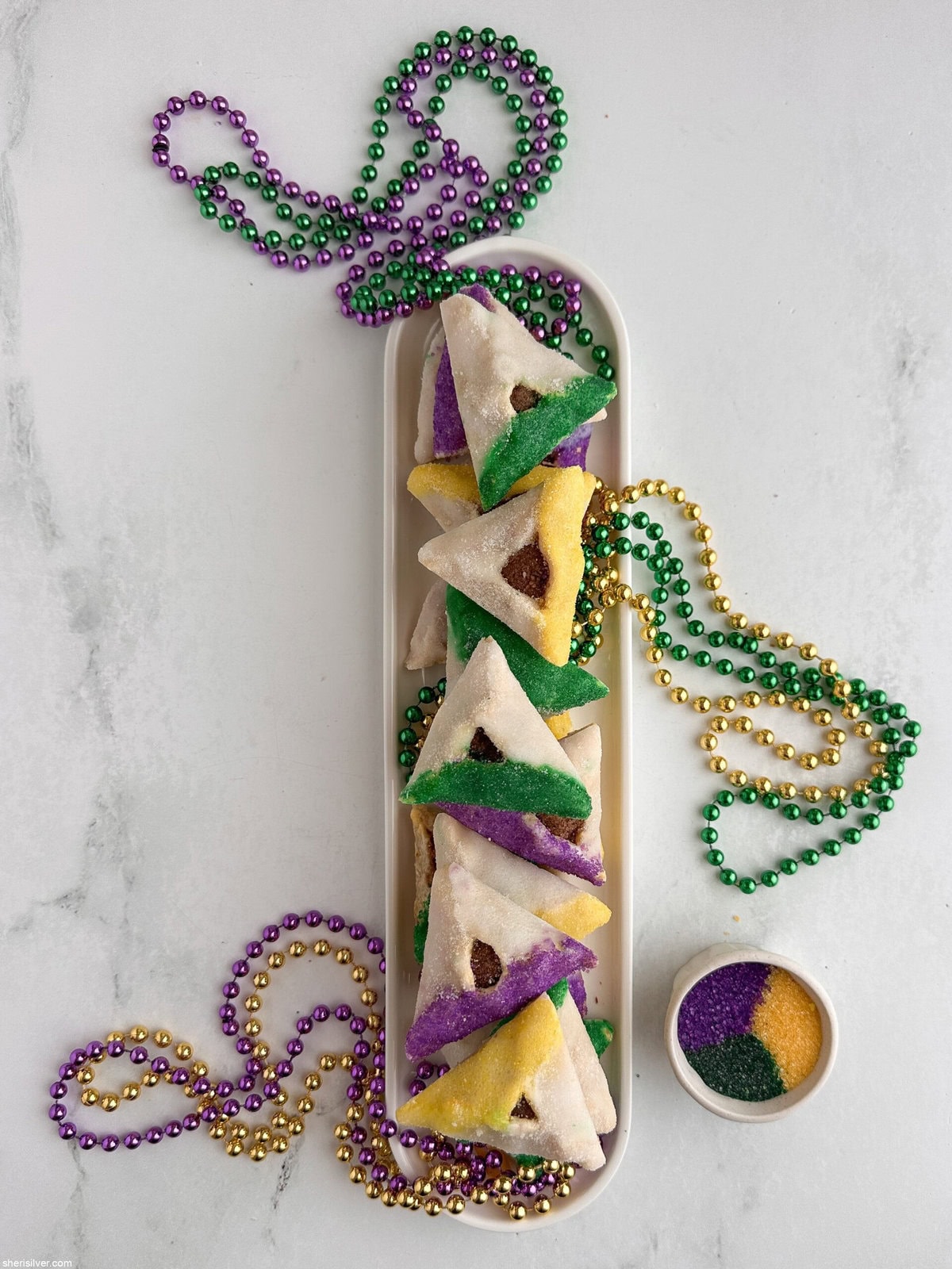King Cake hamantaschen on a white tray with purple, green and gold sanding sugar and Mardi Gras beads