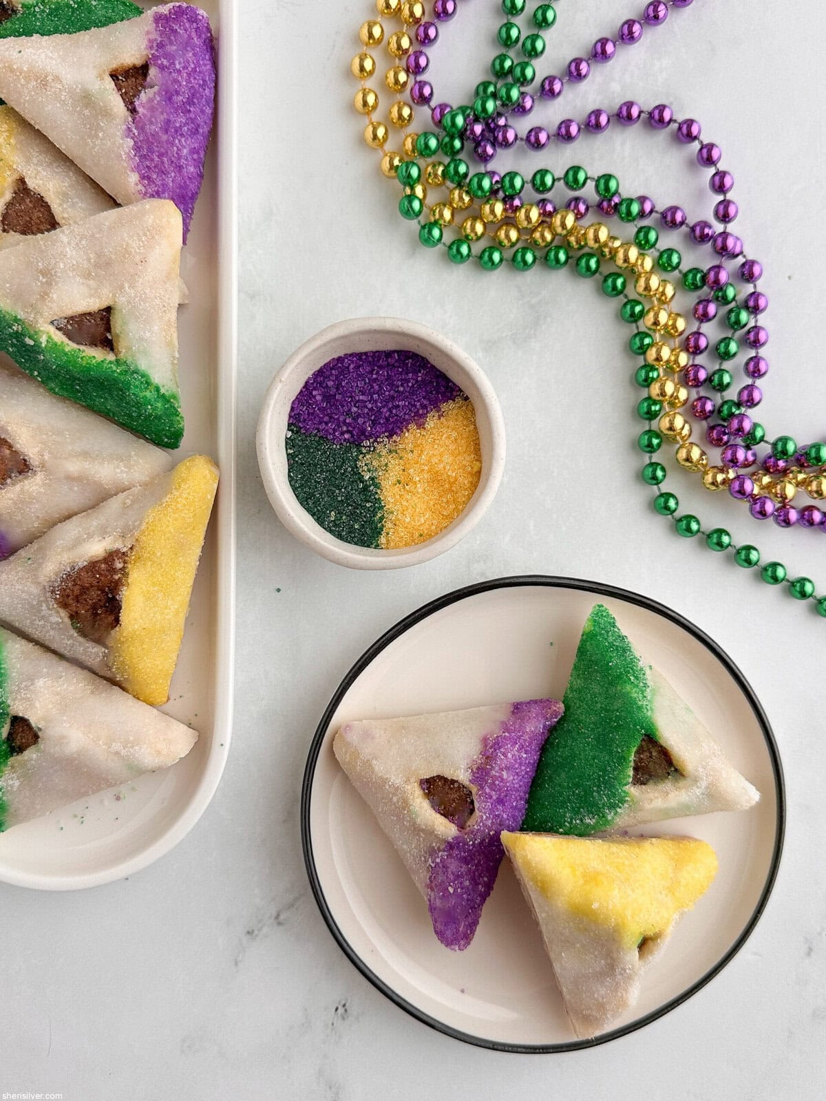 King Cake hamantaschen on a white tray with a small plate showing one cookie in each Mardi Gras color