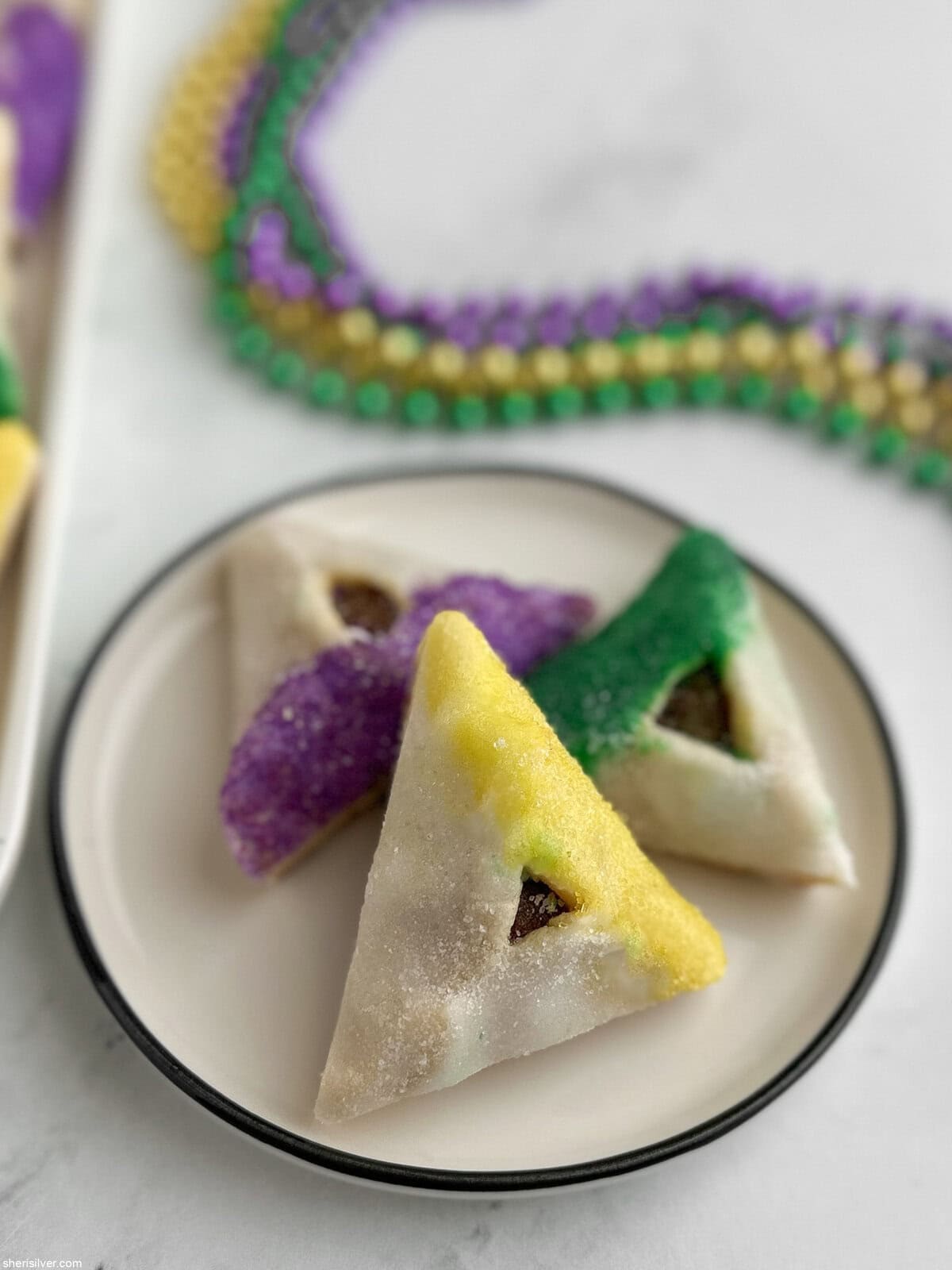 Three King Cake hamantaschen on a small plate with Mardi Gras beads in the background