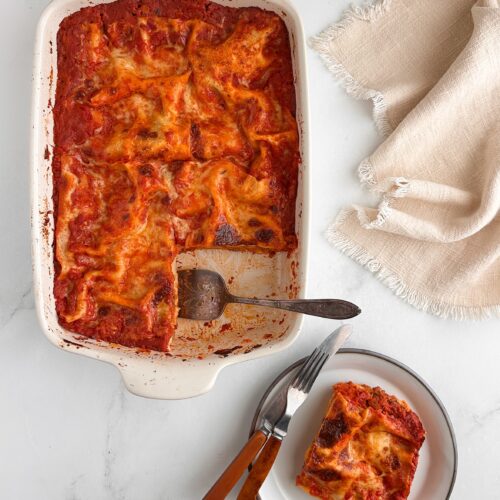 Baked lasagna in a white baking dish with one slice removed and served on a plate.