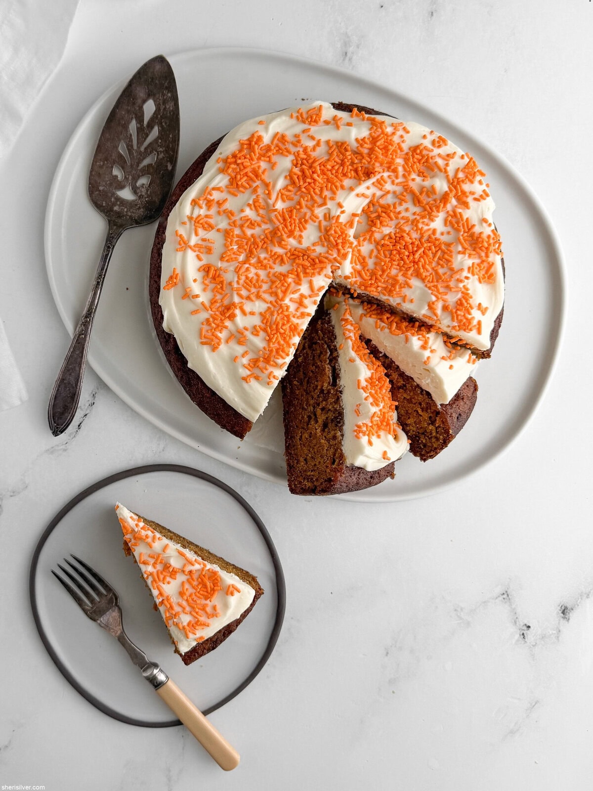Frosted carrot snacking cake with cream cheese frosting and orange sprinkles on a white platter; some slices removed, one slice on a plate