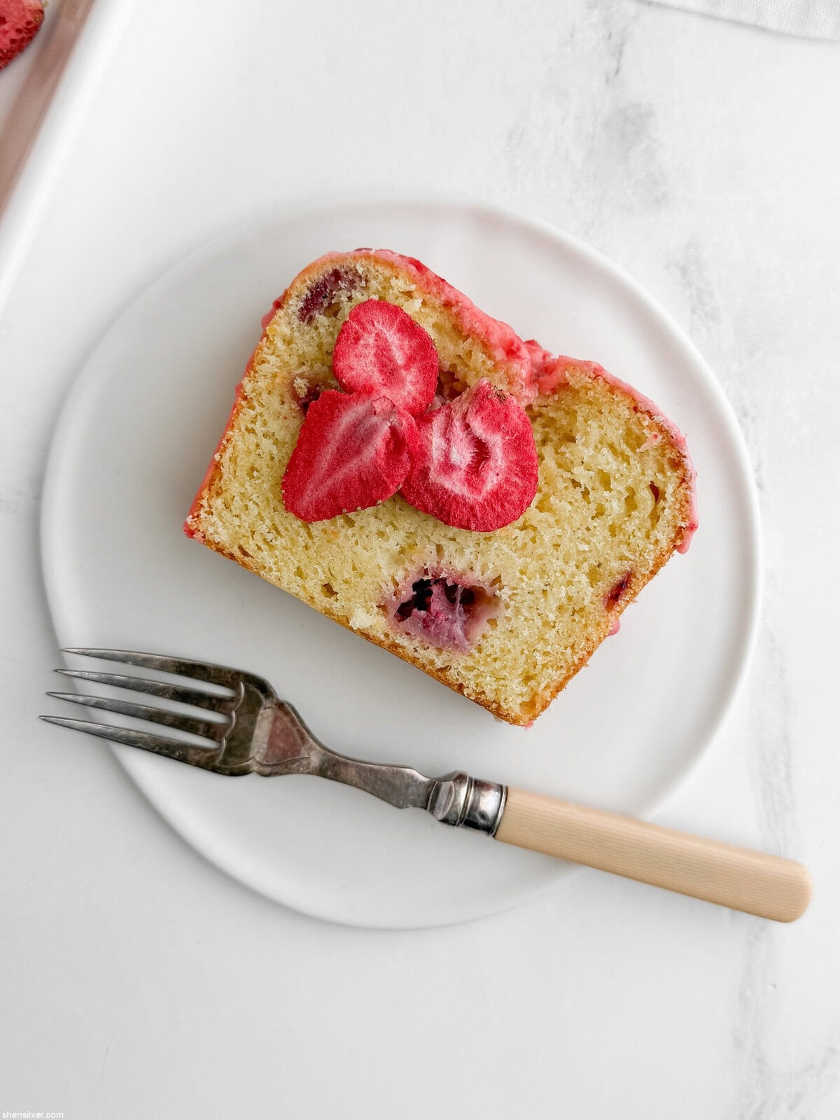 Single slice of strawberry lemon loaf cake on a white plate showing moist crumb, jammy strawberry pockets, pink glaze and freeze-dried strawberry garnish