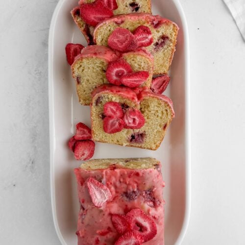 Overhead view of sliced strawberry lemon loaf cake on a white oval platter with pink glaze and freeze-dried strawberry garnish