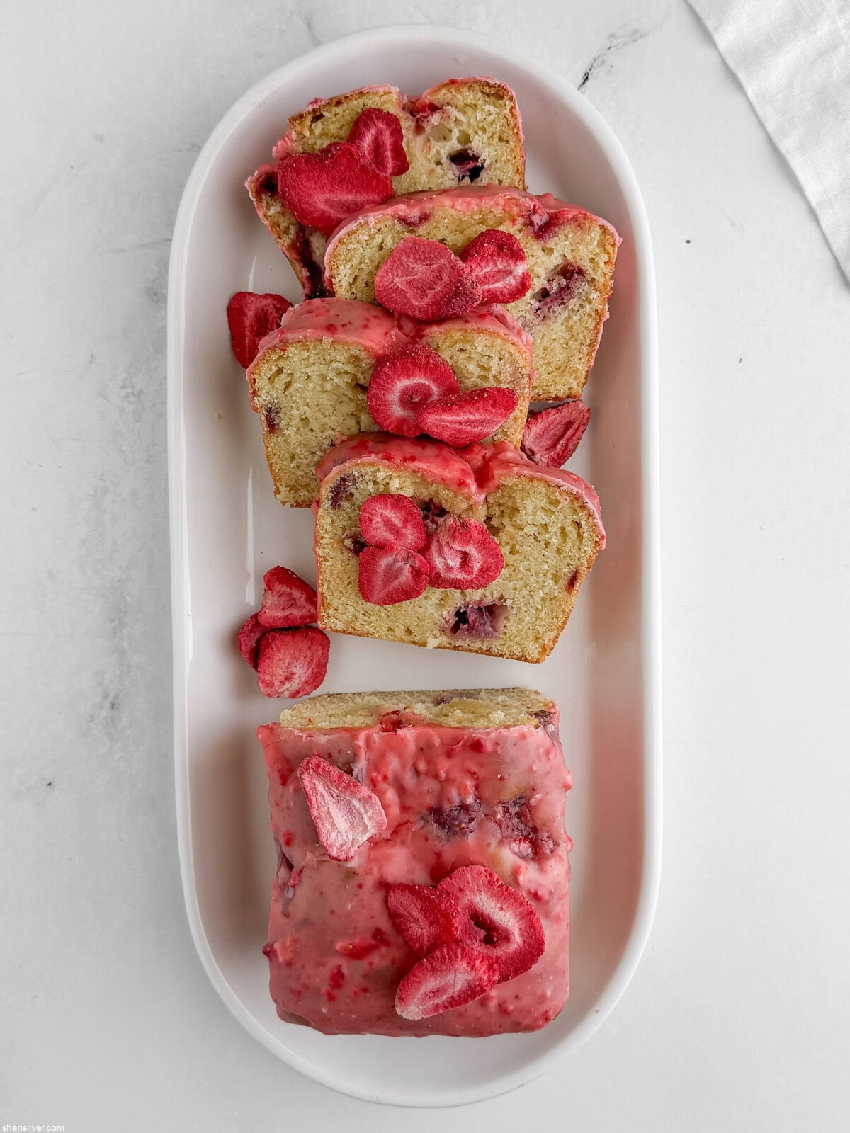 Overhead view of sliced strawberry lemon loaf cake on a white oval platter with pink glaze and freeze-dried strawberry garnish